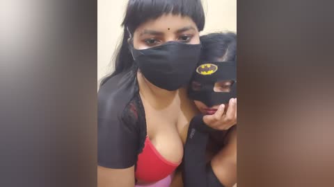 A video of a young South Asian woman with medium skin tone, wearing a black Batman mask, red bra, and black top, capturing a selfie in a bathroom mirror.