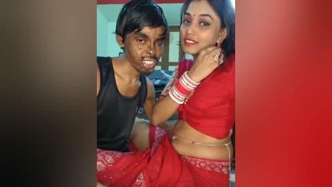 Video of a South Asian couple with dark skin tones, the man in a black tank top and the woman in a red sari, both smiling. Background shows a room with neutral tones and a window.