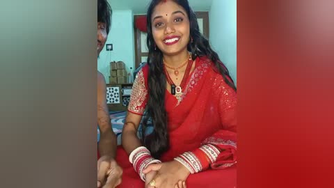 Video of a smiling South Asian woman in red saree with gold embroidery, wearing gold jewelry and red bangles, seated on a bed in a simple, brightly lit room.