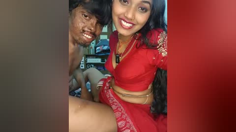 Video of a young Indian woman with long black hair, smiling, in a red saree with gold embroidery, sitting next to a young boy with dark skin, smiling, wearing a red shirt.
