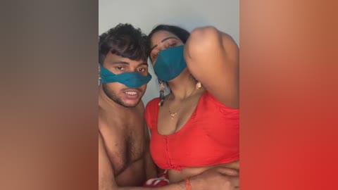 Video of a shirtless South Asian man with curly hair and a mask, standing beside a woman in a red crop top, both smiling, in a dimly lit room.