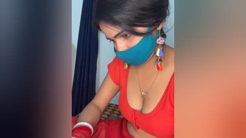 Video of a South Asian woman with medium skin tone, wearing a blue mask, red top, and colorful earrings, seated on a bed, blurred background.