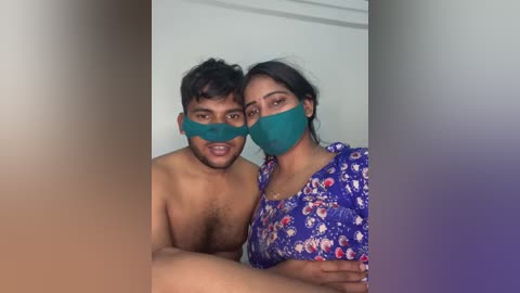 Video of a shirtless South Asian man with short hair and a medium build, and a South Asian woman with long black hair in a floral dress, both wearing green masks, indoors with blurred background.