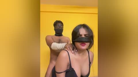 Video of a blindfolded Asian woman in a black bra, being held by a shirtless Black man in a yellow room.