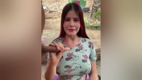 Video of a young woman with straight brown hair, wearing a light blue shirt adorned with cartoon grapes. She is holding hands with another person's hand, partially cropped. Background shows a garden with stone walls and greenery.