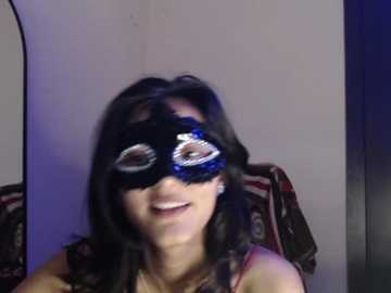 Video of a young woman with light skin and long dark hair, wearing a black and blue masquerade mask, seated on a red-patterned couch.