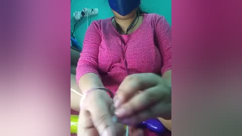 Video of a woman with medium skin tone, wearing a blue mask and pink knit top, manipulating a clear plastic bottle in a teal room.