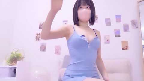 Video of a young Asian woman with a short black bob, wearing a light blue dress with a zipper, posing in a room with white walls and a potted plant.