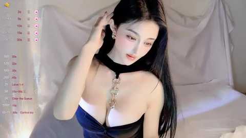 Video of an Asian woman with long black hair, wearing a revealing blue dress with a chain necklace, sitting in a dimly lit room with white drapes.