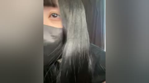 A video shows a person with long, straight, silver hair partially obscuring their face, wearing a black face mask and a black jacket. The background is blurry, focusing on the hair.