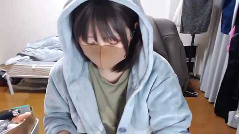 Video of a young Asian woman with shoulder-length black hair, wearing a light blue hoodie and beige face mask, sitting on a wooden floor in a cluttered room with a bed and clothes.