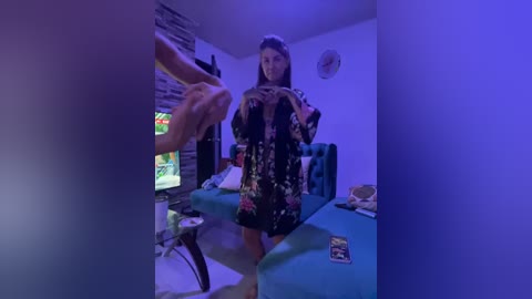 Video of a young woman with long hair, wearing a floral dress, taking a selfie in a dimly lit, cluttered room with a TV and gaming console visible.