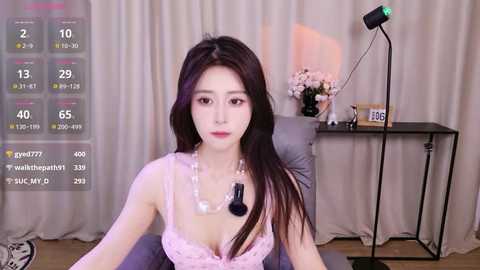 Video of an East Asian woman with long black hair, wearing a pink lace bralette, seated indoors with a monitor showing social media metrics in the background.