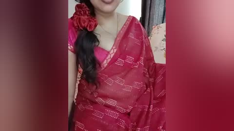 Video of a woman in a red saree with floral patterns, wearing a red flower in her hair, smiling. Background shows a blurred indoor setting with curtains and a wall.