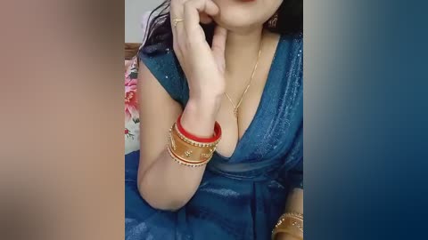Video of a woman with medium skin tone, wearing a blue, sequined sari with a deep V-neck, adorned with gold bangles and a gold necklace. Her long black hair is partially visible, and she is seated indoors, with a blurred floral background.