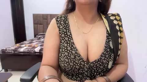Video of a South Asian woman with medium brown skin, large breasts, wearing a low-cut black dress with white floral patterns, a gold necklace, and a black and yellow polka-dot shawl, seated in a bedroom with a bed in the background.