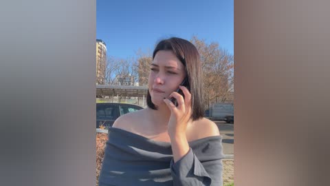 Video of a young woman with shoulder-length dark hair, wearing a loose gray off-shoulder top, speaking on a mobile phone. Background features a clear blue sky, autumn trees, and parked cars.