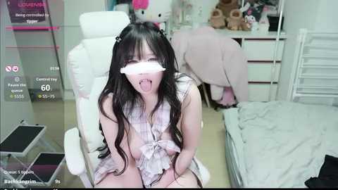 A video of a young, slender Asian woman with long black hair, wearing a pink plaid dress, sitting on a white chair, blindfolded, in a bedroom with stuffed animals, a bed, and a desk.
