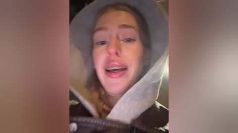 Video of a woman with light skin, light brown hair, and a grey hat, seen from the side. She has a slightly open mouth, suggesting she's talking or singing. The background is blurred, focusing on her face.