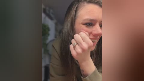 Video of a young woman with light skin and long, straight brown hair, wearing a green jacket, crying, with her fingers on her cheek, in a blurred, indoor background.