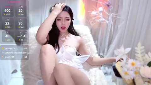 A video of an East Asian woman with long black hair and fair skin, dressed in a white lace lingerie, seated on a fluffy white chair, holding a bouquet of white flowers.