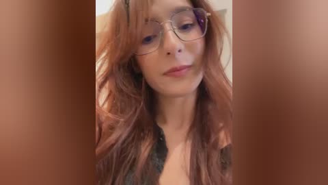 Video of a young woman with long, wavy, auburn hair, wearing round glasses and a black lace top. She has fair skin and a soft smile, with blurred brown background.
