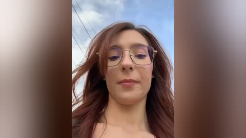Video of a young woman with long, wavy auburn hair, wearing rectangular glasses and a light-colored top, set against a cloudy blue sky background.