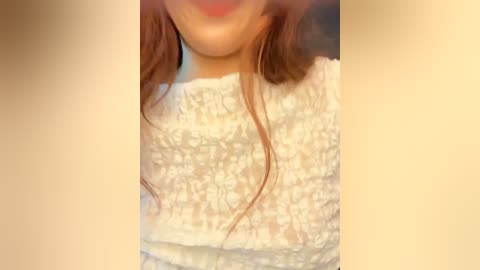 Video of a woman's upper torso in a close-up, showing her wearing a white lace top with intricate patterns. Her long, brown hair cascades down her shoulders. The background is blurred and beige.