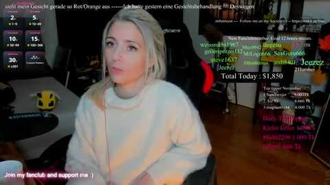 Video of a young woman with blonde hair and fair skin, wearing a white hoodie, seated in a dark, cluttered room with a large monitor displaying chat messages and streaming details.
