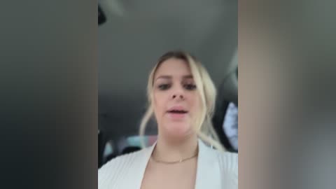 A blurry video of a young woman with light skin and blonde hair, wearing a white blouse, speaking with an open mouth, possibly in a car.