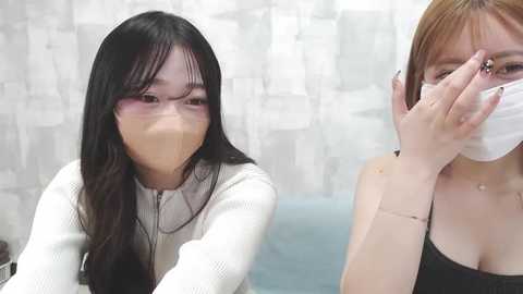 Media: Video of two Asian women with long black hair, wearing face masks, one in a white sweater, the other in a black top, in a modern bathroom with white, abstract-patterned wallpaper.