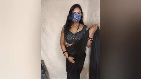 Media: Video of a South Asian woman with medium skin tone, wearing a blue face mask, black saree, and gold jewelry, holding a black garment, in a plain, beige-walled room.