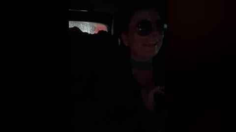 A dimly lit video shows a young woman with short, dark hair and sunglasses, wearing a striped shirt, peeking out from a darkened window. The background is dark and indistinct.