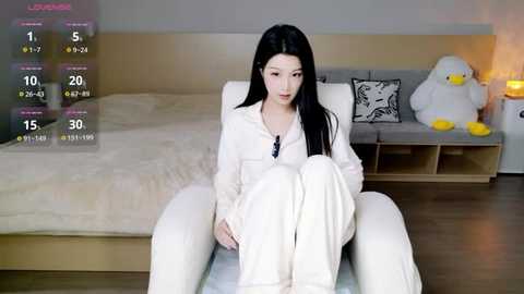 A video of an East Asian woman with long black hair, wearing white pajamas, sitting in a white armchair in a minimalist bedroom with a wooden floor and a gray cushioned bed.