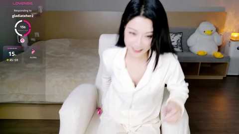 Video of a young Asian woman in a white robe, sitting on a white couch, with a plush duck and a lamp in the background.