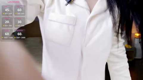 Video of a woman wearing a white zip-up jacket with pockets, standing indoors. The background features dim lighting and indistinct furniture. The image includes a virtual overlay with temperature and weather information.