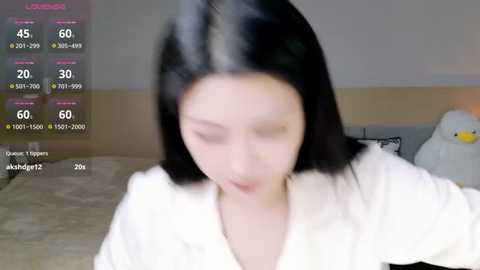 Video of a young East Asian woman with straight black hair, wearing a white robe, sitting in a dimly lit bedroom with a plush duck toy and a bed in the background.