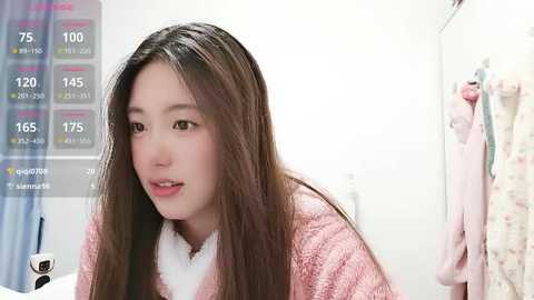 A video of a young Asian woman with long, straight brown hair, wearing a pink, fuzzy robe, in a white-tiled bathroom with a hanging towel and a digital thermometer display.