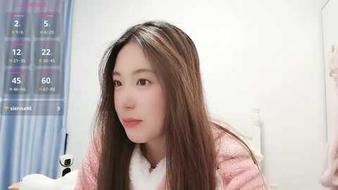 A video of a young East Asian woman with long, straight brown hair, fair skin, and light makeup, wearing a pink quilted jacket, sitting in a medical examination room with a blue and white calendar on the wall and a stethoscope in her hand.