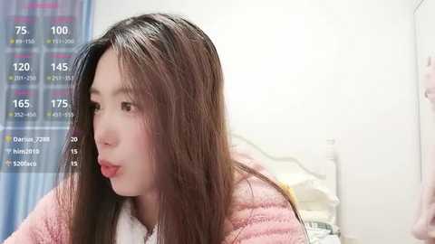 A video of a young Asian woman with straight, long brown hair, wearing a pink sweater, looking pensive, in a bright, modern bedroom with a white wall and a colorful digital display.