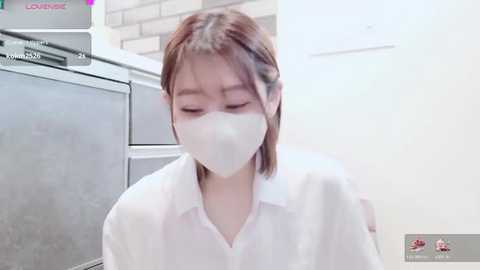 Video of an East Asian woman with light skin and shoulder-length brown hair, wearing a white mask and shirt, sitting on a grey laundry machine in a modern, clean laundry room.