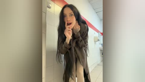 Video of a slender, pale-skinned woman with long black hair, wearing a dark brown coat, in a modern, brightly lit restroom with white tiles and red accents. She playfully covers her mouth with her hand.