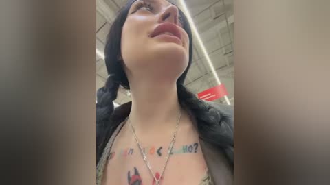 Media: A close-up video of a young woman with fair skin, wearing a black beanie, glasses, and a jacket, displaying multiple tattoos on her chest. She stands in an indoor setting with exposed concrete ceiling.