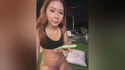 Video of a young, slender woman with long, auburn hair, wearing a black sports bra and white shorts, holding a green cucumber, seated outdoors on a grassy lawn.