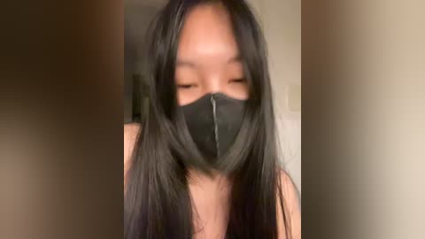 A close-up video of a young Asian woman with long black hair, wearing a black face mask and a sleeveless top, indoors with blurred background.