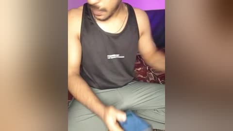 Video of a fit, light-skinned man with a trimmed beard, wearing a black tank top and green pants, sitting on a patterned couch, holding a blue object.