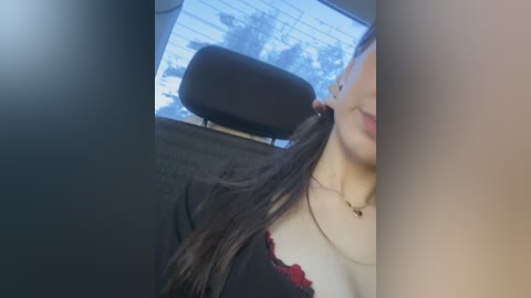 Video of a woman with long dark hair, wearing a black lace-trimmed top, sitting in a car, partially visible through the window, with blurred outdoor scene.