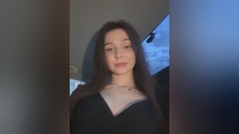 Video of a young woman with long dark hair, fair skin, and a black dress, smiling. The background shows a dark room with a window and a snow-covered ground.