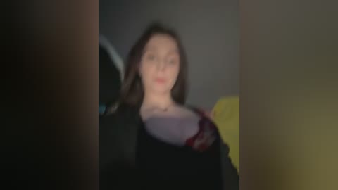 A blurry, dimly-lit video of a woman with fair skin, dark hair, and a black top, standing indoors, partially obscured by a yellow object and a dark figure in the foreground.