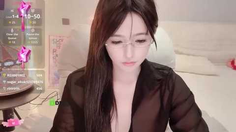 Video of an Asian woman with fair skin, long black hair, and glasses, wearing a sheer black top, sitting indoors. The background includes a bed, pink flowers, and a digital display.